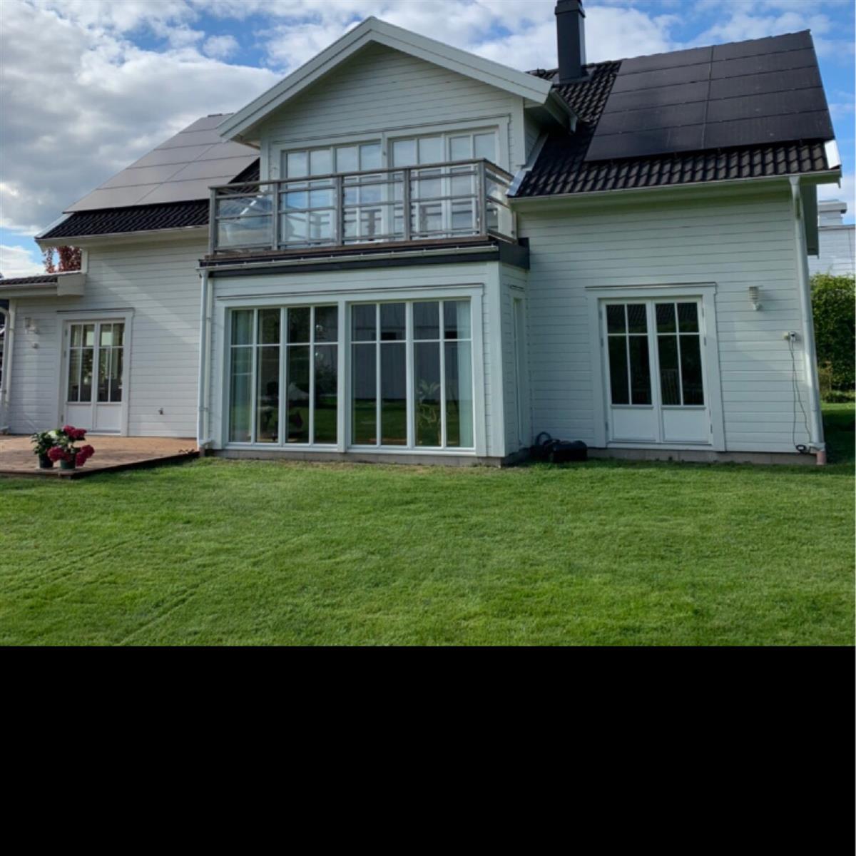 Ett tvåvånings vitt hus med stora fönster, solpaneler på taket, en glasbalkong och en välskött gräsmatta under en halvmolnig himmel.