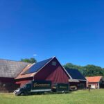 Röd ladugård med solpaneler på taket, omgiven av gräs och träd. En lastbil och släp med Solpanelen.nu står parkerade i närheten under en klarblå himmel.