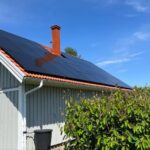 Solpaneler på ett rött tegeltak i ett ljusgrått hus med skorsten, omgivet av gröna buskar, under en klarblå himmel.