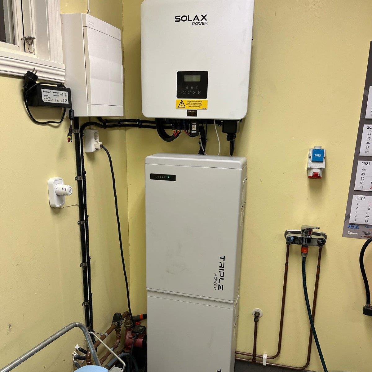 Batterilagringssystem märkt SolaX Power och en Triple Power-enhet monterad på en gul vägg i ett grovkök, med en kalender och olika kablar i närheten.
